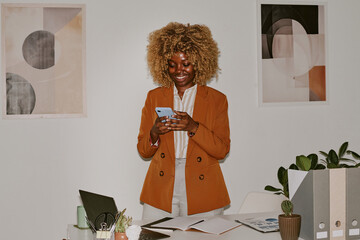 Blonde curly haired woman texting in social network and smiling widely while standing next to her...