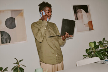 Dutch angle shot of busy young influencer holding laptop while speaking on phone