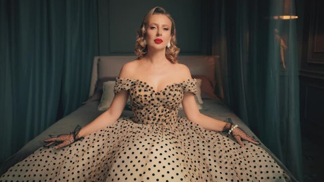 Blonde hair retro lady beauty woman pinup posing sitting on bed in luxury bedroom. old-fashioned Vintage polka dot white black beige dress. Girl fashion model red lips sexy eyes 30s pin-up old style
