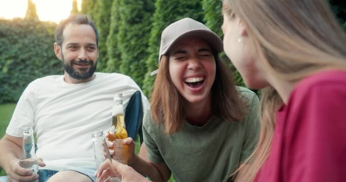Happy brunette girl in a green t-shirt and cap chats with her friends and drinks beer while spending time in the yard and having a picnic in the summer