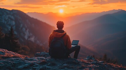 Obraz premium A lone hiker sits on a mountaintop at sunset, working on a laptop.