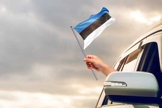 Waving the Estonia flag against the sunrise or sunset from a car driving along a country road. Holding the Estonia flag, traveling by car, on a weekend trip. - Powered by Adobe