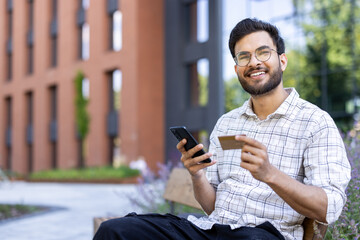 Portrait of a young Indian man holding a credit card and a mobile phone, sitting on a bench outside...