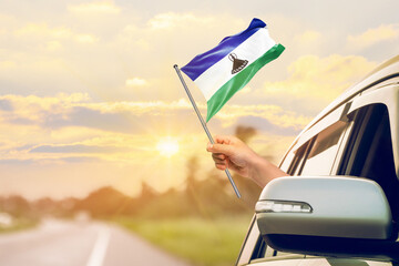 Waving the Lesotho flag against the sunrise or sunset from a car driving along a country road. Holding the Lesotho flag, traveling by car, on a weekend trip.