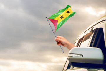 Waving the S&atilde;o Tom&eacute; and Pr&iacute;ncipe flag against the sunrise or sunset from a car driving along a country road. Holding the S&atilde;o Tom&eacute; and Pr&iacute;ncipe flag, traveling by car, on a weekend trip.