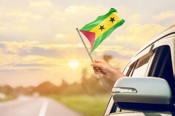 Waving the S&atilde;o Tom&eacute; and Pr&iacute;ncipe flag against the sunrise or sunset from a car driving along a country road. Holding the S&atilde;o Tom&eacute; and Pr&iacute;ncipe flag, traveling by car, on a weekend trip.