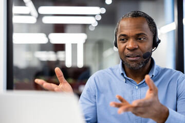 Confident professional man wearing headset in modern office engaged in video conference. Gesturing with hands, focused expression indicating thoughtful communication with team