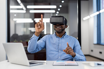 Businessman wearing virtual reality headset engaged with virtual environment using hand gestures. Seated at office desk, smiling, utilizing laptop and book for work. Innovation and technology