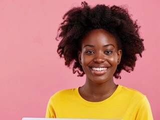 A woman with curly hair is smiling and wearing a yellow shirt. She is holding a laptop in her hand