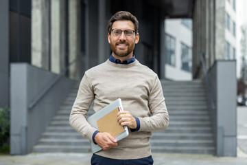 Businessman standing outside office building holding laptop. Smiling confidently, wearing glasses and casual clothing. Represents modern professional, ready for work.