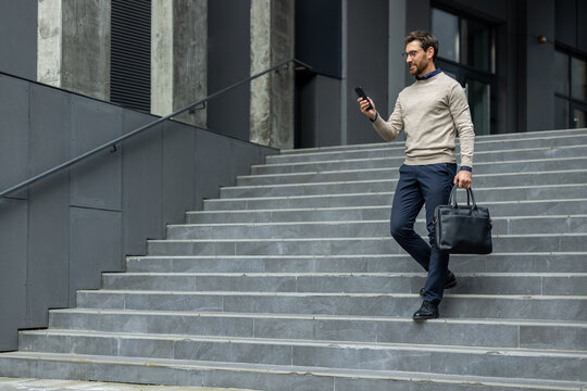 Businessman walking down grey concrete steps using smartphone, dressed in smart casual outfit, holding black leather briefcase. Image represents professional urban lifestyle, confidence, communication - Powered by Adobe