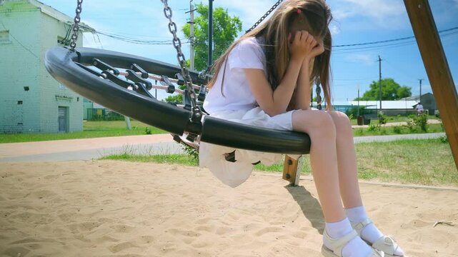 Upset ittle girl kid swinging on net swing in the park. Caucasian child swinging on playground. Cute child have saddnes in park. School girl play on swing. Slow motion
