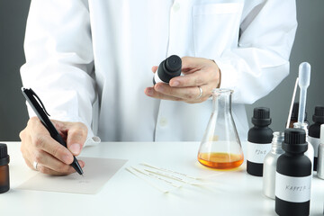 right hand is writing testing result of blending scents for making perfume and candle by perfumer in laboratory with chemical beaker blotting paper , fragrance bottle and essential oil bottle on table