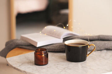 Cup of coffee with scented candle and open paper book on marble table in room close up. Cozy home atmosphere.