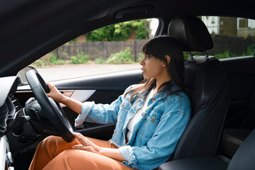 Young Woman in Denim Jacket Focused on Driving Through a Serene Neighborhood in Daylight