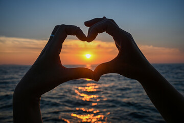 Woman making heart shape with hands framing setting sun over the ocean