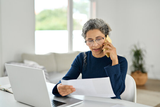 Busy middle aged woman working from home making phone call checking document. Mature older lady having mobile conversation talking on the phone while hoding paper calling customer support service.