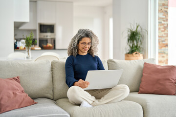 Happy curly mature older woman using laptop sitting on couch at home. Smiling middle aged lady...