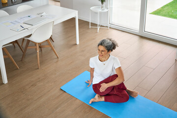 Happy healthy fit mature woman practicing yoga doing exercises sitting on mat at home interior....