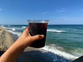 Hand holds up a soda pop in a plastic cup, with a view of the ocean. Taken in the Outer Banks of North Carolina