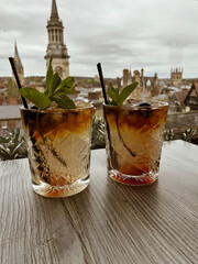 Two refreshing cocktails sit on a table in Oxford, overlooking the historic city skyline with iconic buildings in the background.