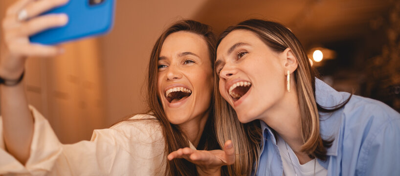 Two young beautiful smiling hipster sisters friends female in trendy summer clothes. Carefree women rest sitting in cafe restaurant. Positive models having fun and taking selfie on smartphone. 