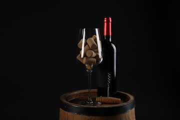 Corkscrew, bottle of wine, glass and corks on wooden barrel against black background