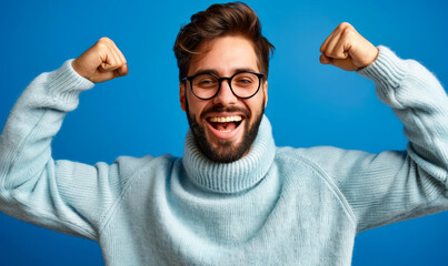 Naklejka premium Excited Young Man Celebrates Success with Arms Raised, Beaming with Joy - Casual Sweater, Glasses, Beard, Blue Background - Winning, Achievement