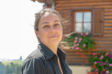 Street portrait of a dark-haired joyful woman 40-45 years old on the background of a wooden house with flowers.