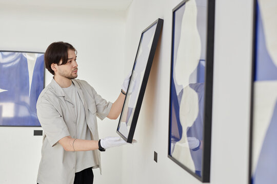 Minimal side view portrait of young Asian man working as art curator and hanging paintings on wall while planning modern art gallery exhibition copy space