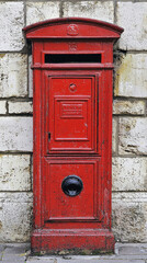 A vintage red British postbox with a black handle and a small opening for mail. The postbox is mounted on a brick wall.