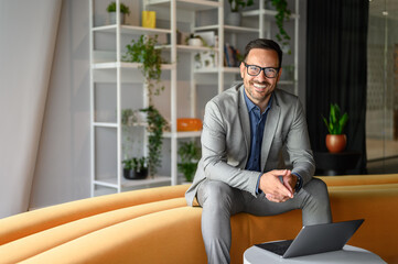 Portrait of young contented businessman in glasses sitting with laptop on yellow seat in modern office