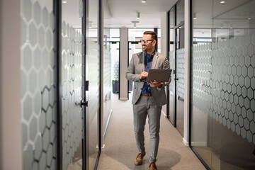 Full length of male professional working over laptop and looking away confidently in office corridor