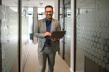 Confident male professional working over laptop and smiling at camera while standing in office corridor
