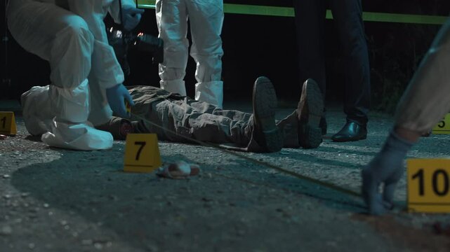 Detective and forensic scientists working hard at crime scene near headless body. Collection and photography of evidence, measurements, examination of the victim's body. Night scene.