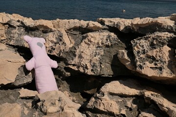 Cute teddy bear handmade of white fabric with think pink vertical lines standing on porous shoreline rocks, deep blue bay water visible in background. Location south of Razanac town, Croatia. 