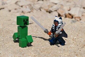 Naklejka premium LEGO Star Wars figure of female Togruta Jedi Ahsoka Tano with two lightsabers ready to fight with green explosive LEGO Minecraft mob Creeper on shoreline rocks, summer daylight sunshine. 