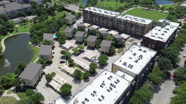 Lakeside urban apartment building complex shingle roofing, flat roof industrial rooftop packaged AC, furnace unit in heavy duty steel cabinet, lush green park pond, trails, Keller Texas, flyover
