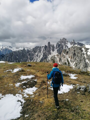 Fototapeta premium A lone hiker wearing a blue jacket, red hat, and carrying trekking poles traverses a grassy area among the snowy peaks of the Dolomites on a cloudy spring day.