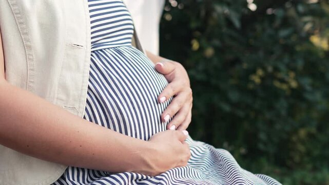 Pregnant woman sitting in summer home garden. Future mom caressing belly bump, green nature background. Expectant mother in summer dress, happy motherhood.
