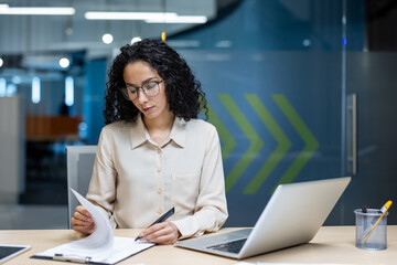 Confident Hispanic businesswoman focused reviewing documents in office setting. Wearing glasses, seated at desk with laptop and paperwork. Represents professionalism, determination, and concentration