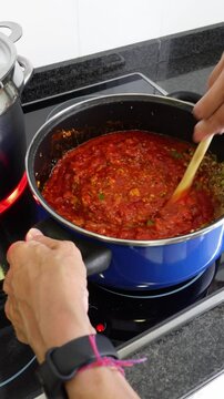 Vertical shot of hands of woman cooking bolognese sauce, mixing fried tomato, minced meat, zucchini, slices of mushrooms and diced onion with a wooden spoon, next to stainless saucepan on an electric 