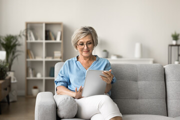 Aged female resting on sofa with modern tablet device, read e-book, articles, or news, checking her...