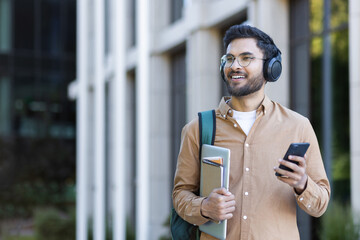 Confident smiling student wearing headphones holds laptop and smartphone outdoors. Casual attire...