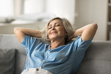 Calm older woman leaning back on sofa with hands behind head, resting with eyes closed, with...