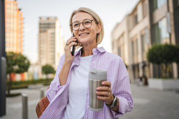 Happy mature woman walk with thermos talk on cellphone on the street