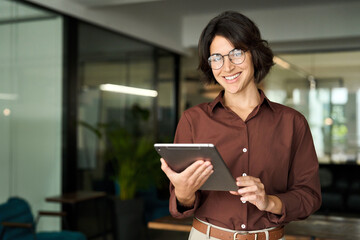 Smiling young Hispanic professional business woman wearing glasses using tablet standing in office....