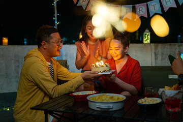 Group of Happy Asian people friends having celebration birthday party together at outdoor rooftop with food and drink. Adult Woman excited with birthday cake and blowing birthday candle with happiness
