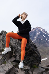 Authentic Caucasian Ethnicity Woman is sitting on rock in mountains, with cone of volcano looming in distance. Millennial woman, enjoying moment of rest, raises hands to head, looks off into distance