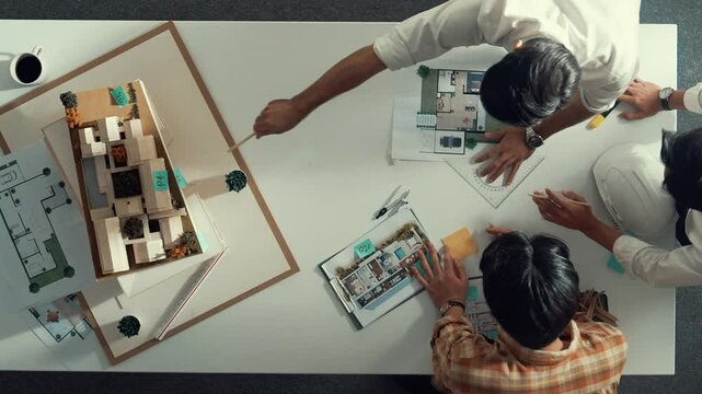 Group of skilled designer looking at model while discussing about building. Top view of architect engineer explain idea while talking and writing blue print scattering at meeting table. Alimentation.